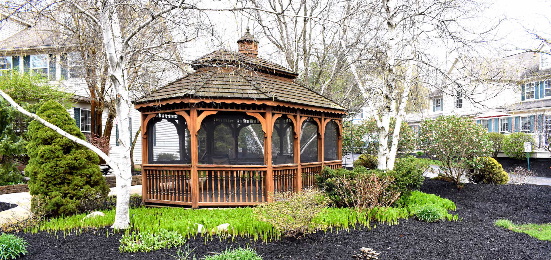 A wooden gazebo with a shingled roof surrounded by landscaped garden beds with green plants, bushes, and trees in front of a multi-story residential building.