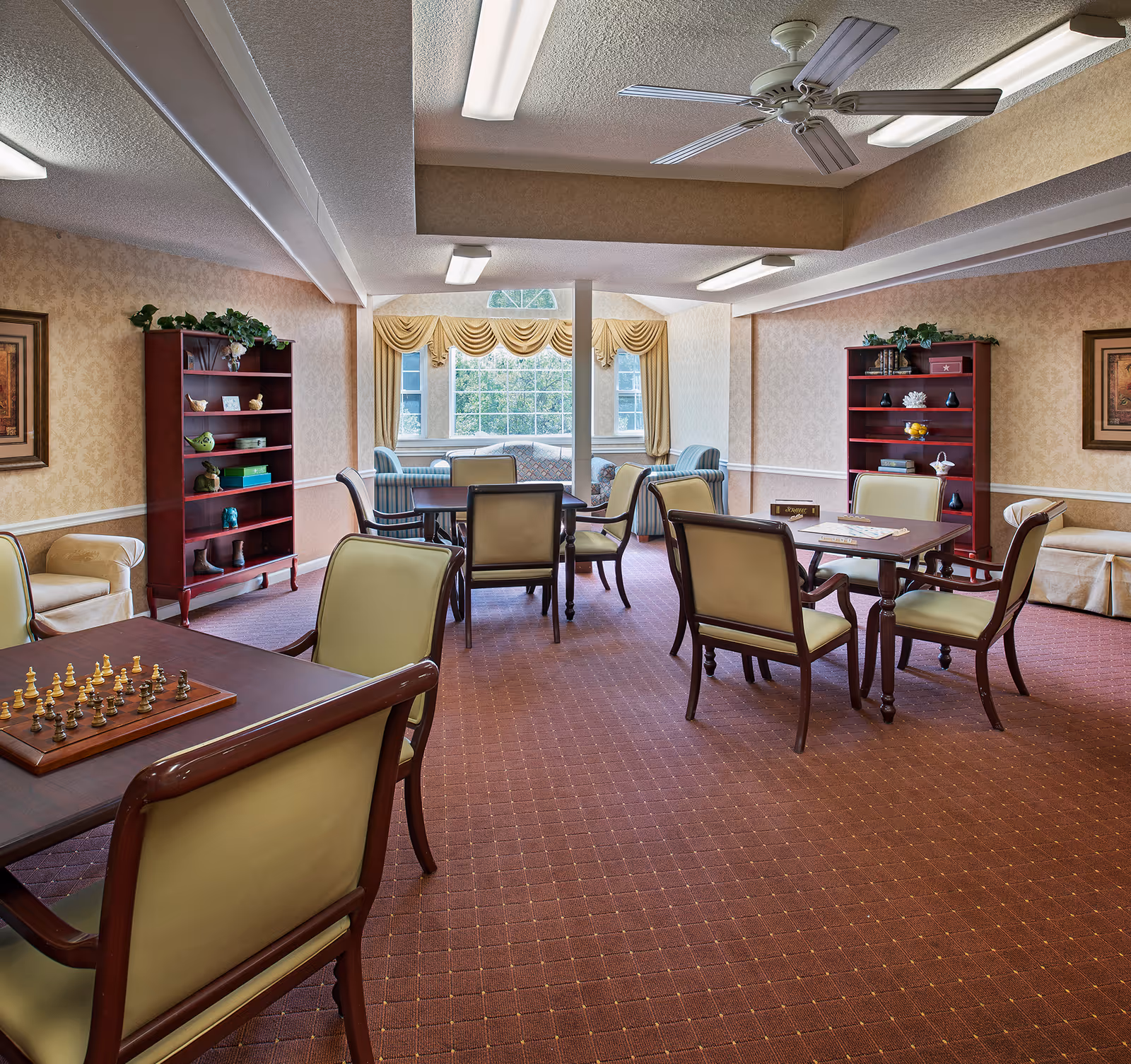 A spacious common room with multiple tables and chairs arranged for games and socializing. One table has a chess set, and another has a Scrabble game. The room features two red bookshelves with decorative items, beige walls with patterned wallpaper, a large window with yellow curtains, and ceiling fans with fluorescent lighting.