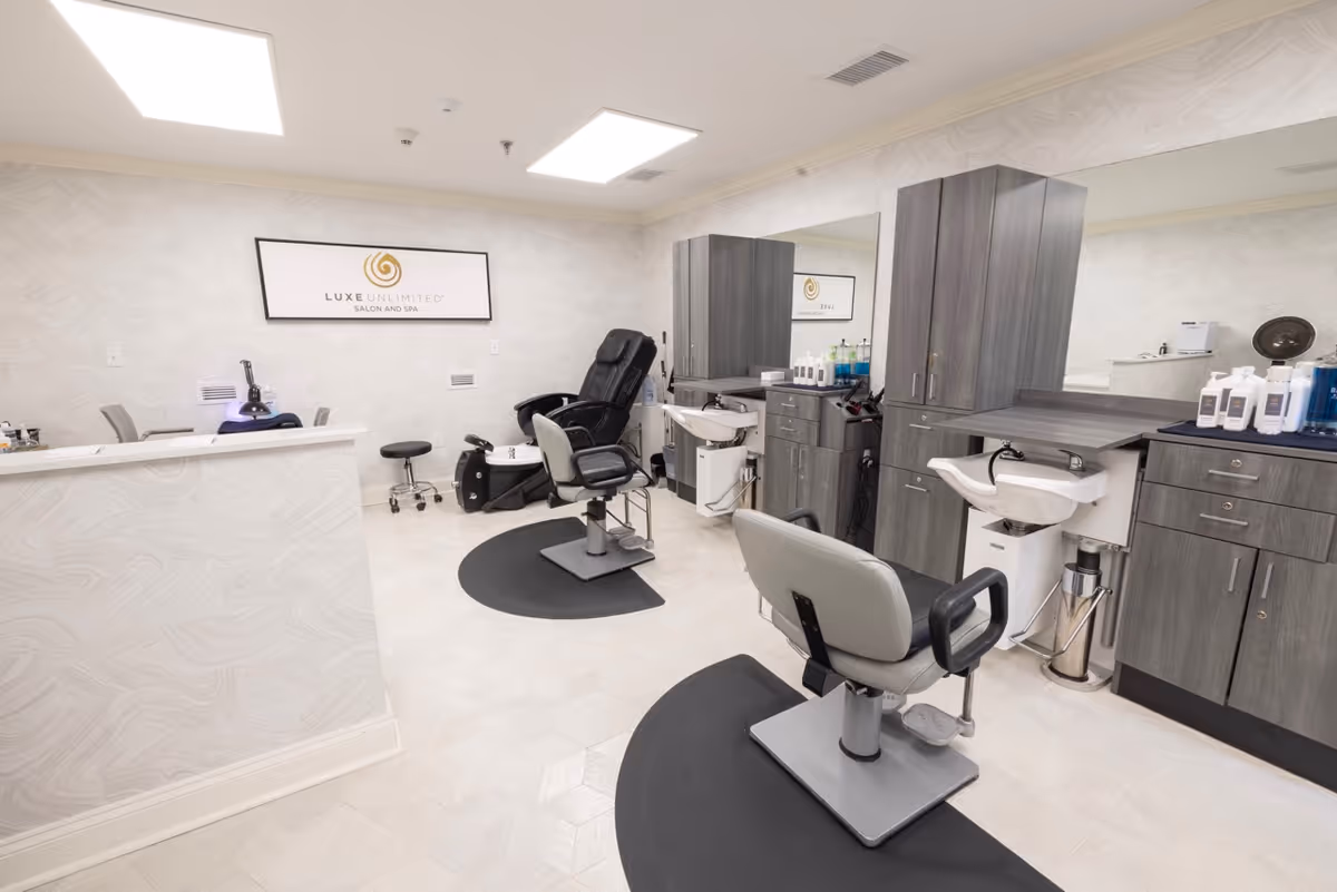 Interior view of a modern salon and spa area with two salon chairs, sinks for hair washing, dark wood cabinetry, and a black reclining chair. The walls are light-colored with subtle patterns, and there is a sign on the wall that reads 'Luxe Unlimited Salon and Spa'. The floor is light-colored with a hexagonal tile pattern.
