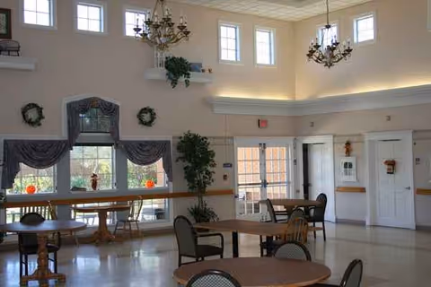Bright senior living common area with round tables, chairs, chandeliers, and large windows.