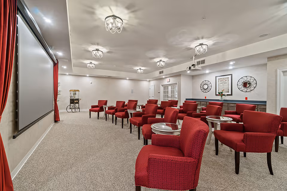 A screening room with rows of red upholstered chairs and small round tables facing a large projection screen under decorative ceiling lights.