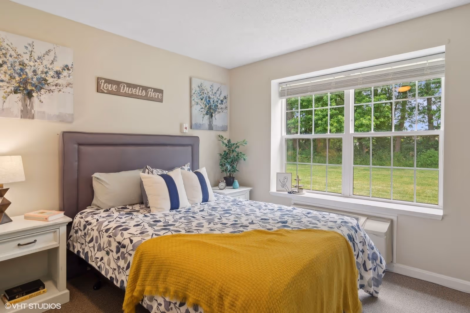 A cozy bedroom with a large window showing a green outdoor view. The bed has a gray upholstered headboard, white and blue floral bedding, and a mustard yellow throw blanket. There are two white nightstands on either side of the bed, one with a lamp and books, and the other with a plant and a clock. Above the bed are two floral paintings and a wooden sign that reads 'Love Dwells Here'.