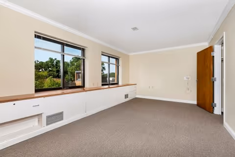 Empty carpeted interior room with beige walls, two large windows overlooking trees, built-in white cabinets under the windows, and an open wooden door.