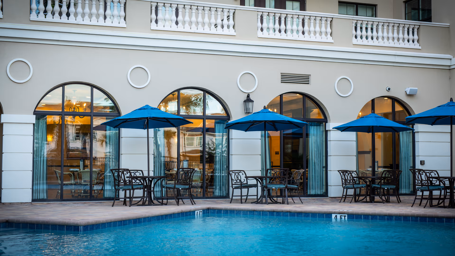 Outdoor pool area with blue umbrellas and black metal tables and chairs in front of a building with large arched windows and white decorative trim.