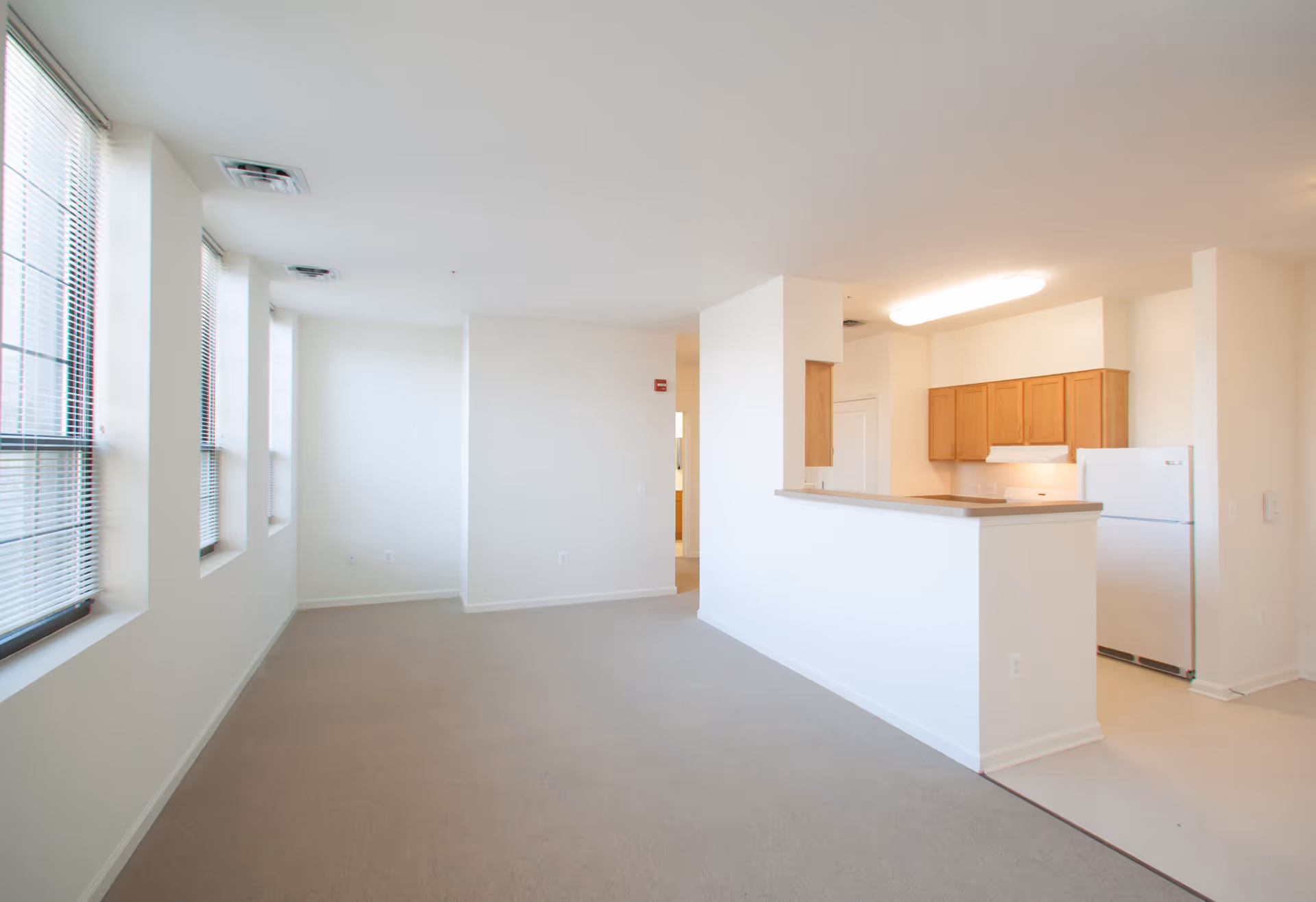 Empty interior space with beige carpet and white walls featuring large windows with blinds on the left side. On the right side, there is a kitchen area with wooden cabinets, a white refrigerator, and a counter with a raised bar separating the kitchen from the main room.