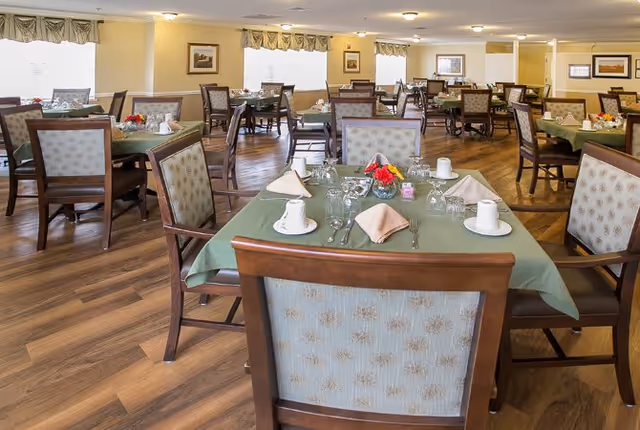A spacious dining room with multiple tables covered in green tablecloths, each set with white cups, napkins, silverware, and small floral centerpieces. The room has wooden flooring, cushioned chairs with patterned upholstery, and large windows with valances allowing natural light to fill the space.