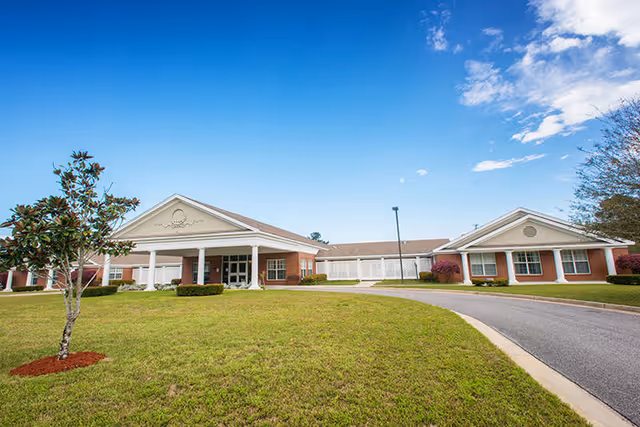 Front exterior view of a single-story brick building with white columns and a covered entrance, surrounded by a well-maintained lawn and a small tree under a blue sky with some clouds.