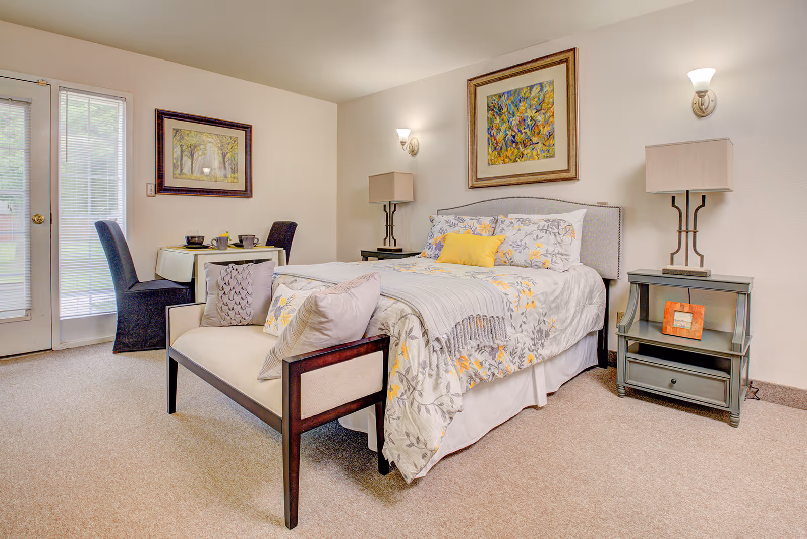 Bright furnished bedroom with a bed, bench at the foot, nightstands, small dining table and a glass patio door.