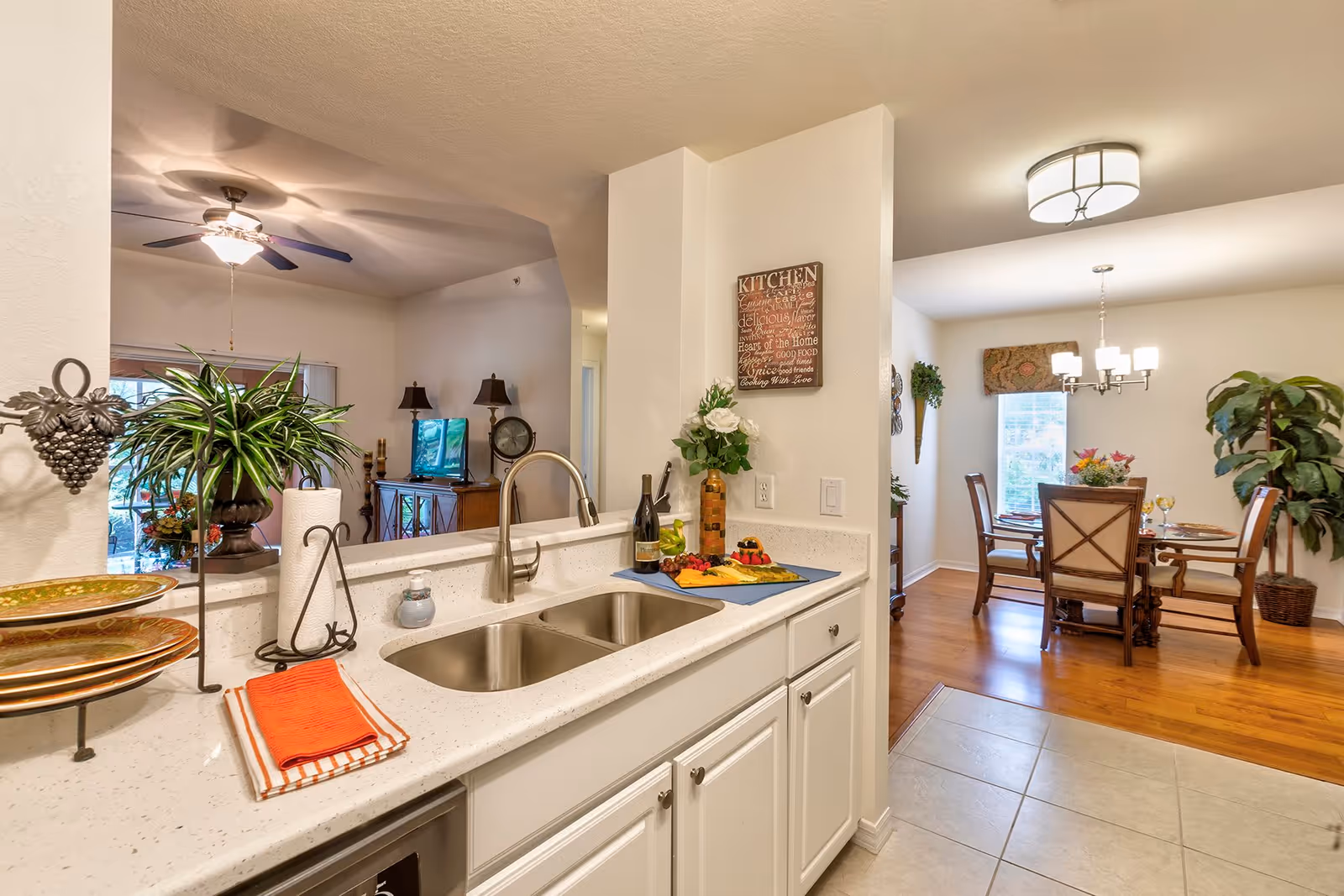 View of a modern kitchen with a double sink, white countertops, and cabinetry. The kitchen opens to a dining area with a wooden dining table and chairs, a chandelier, and large windows letting in natural light. There are decorative plants and a wall sign in the kitchen area. A living room with a ceiling fan and TV is partially visible in the background.