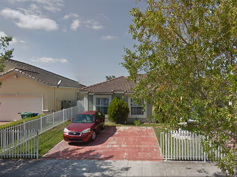 Front exterior view of a single-story house with a red car parked in the driveway, a white picket fence on both sides, and a tree partially obscuring the right side of the house under a partly cloudy sky.