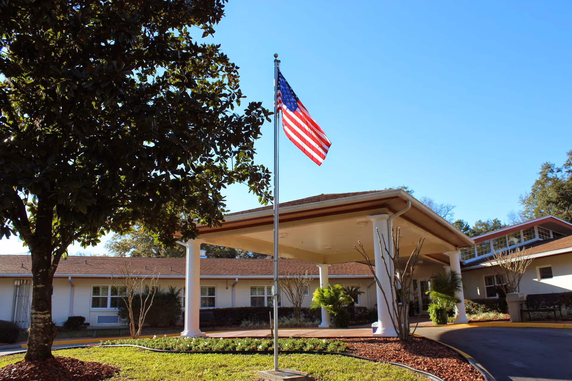 Front entrance of a senior living facility with an American flag on a flagpole, a covered drive-through entry, and landscaped grounds.