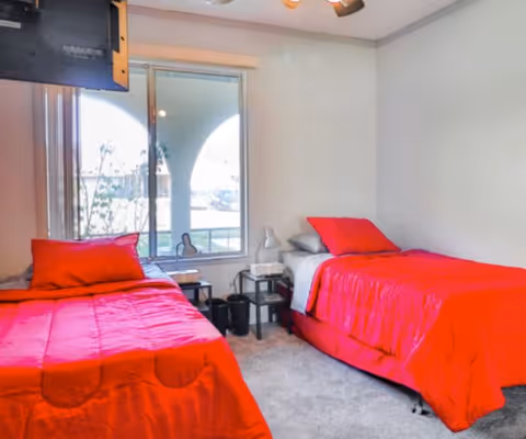 A bright bedroom with two single beds covered in red bedspreads and pillows. There is a window between the beds showing an outdoor view with an archway and some plants. A small black nightstand with a lamp and other items is placed between the beds. A TV is mounted on the wall above the left bed.