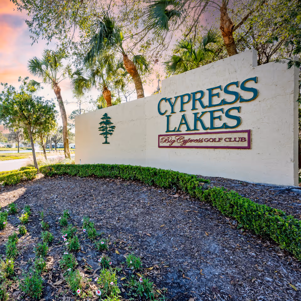Outdoor entrance sign for Cypress Lakes with the text 'Big Cypress Golf Club' below it, surrounded by palm trees and landscaping under a colorful sky at sunset.