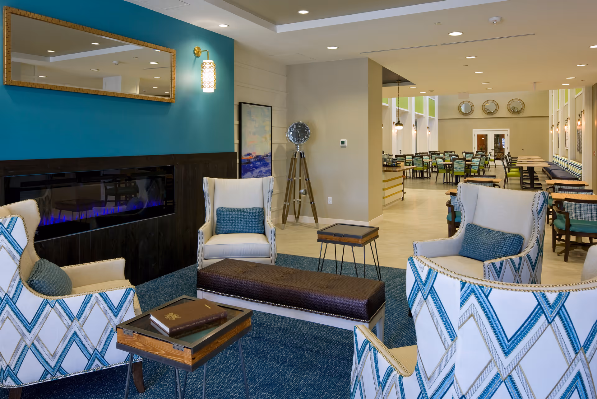 Bright communal lounge with upholstered armchairs around a modern fireplace and a dining area visible in the background.
