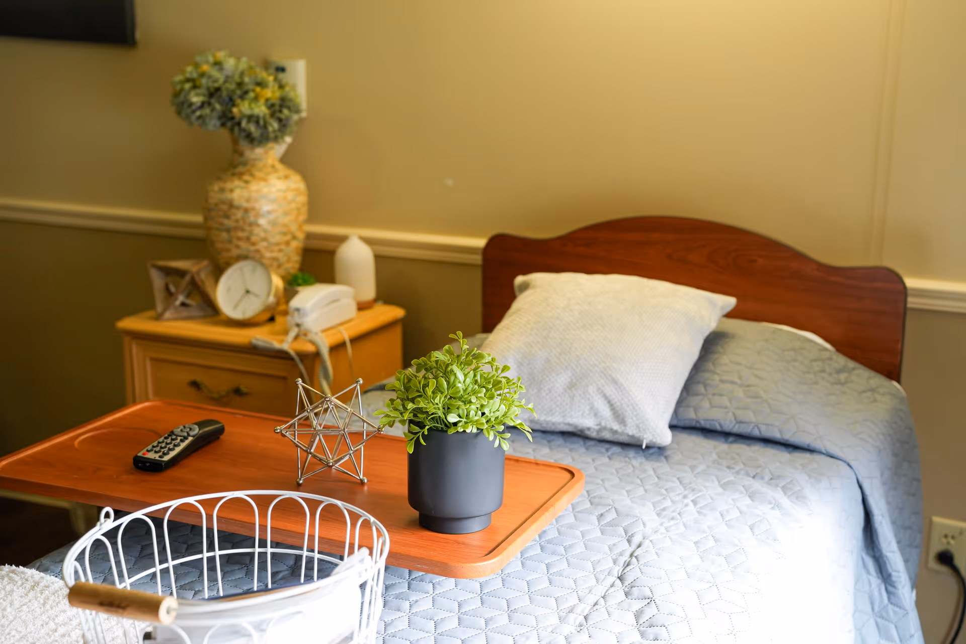 A neatly made single bed with a light blue quilt and a white pillow in a room. A wooden bedside table next to the bed holds a large decorative vase with flowers, a clock, a telephone, and a small white bottle. A wooden tray on the bed holds a small green potted plant, a decorative metal star, and a TV remote control. The room has beige walls and a power outlet is visible near the bed.