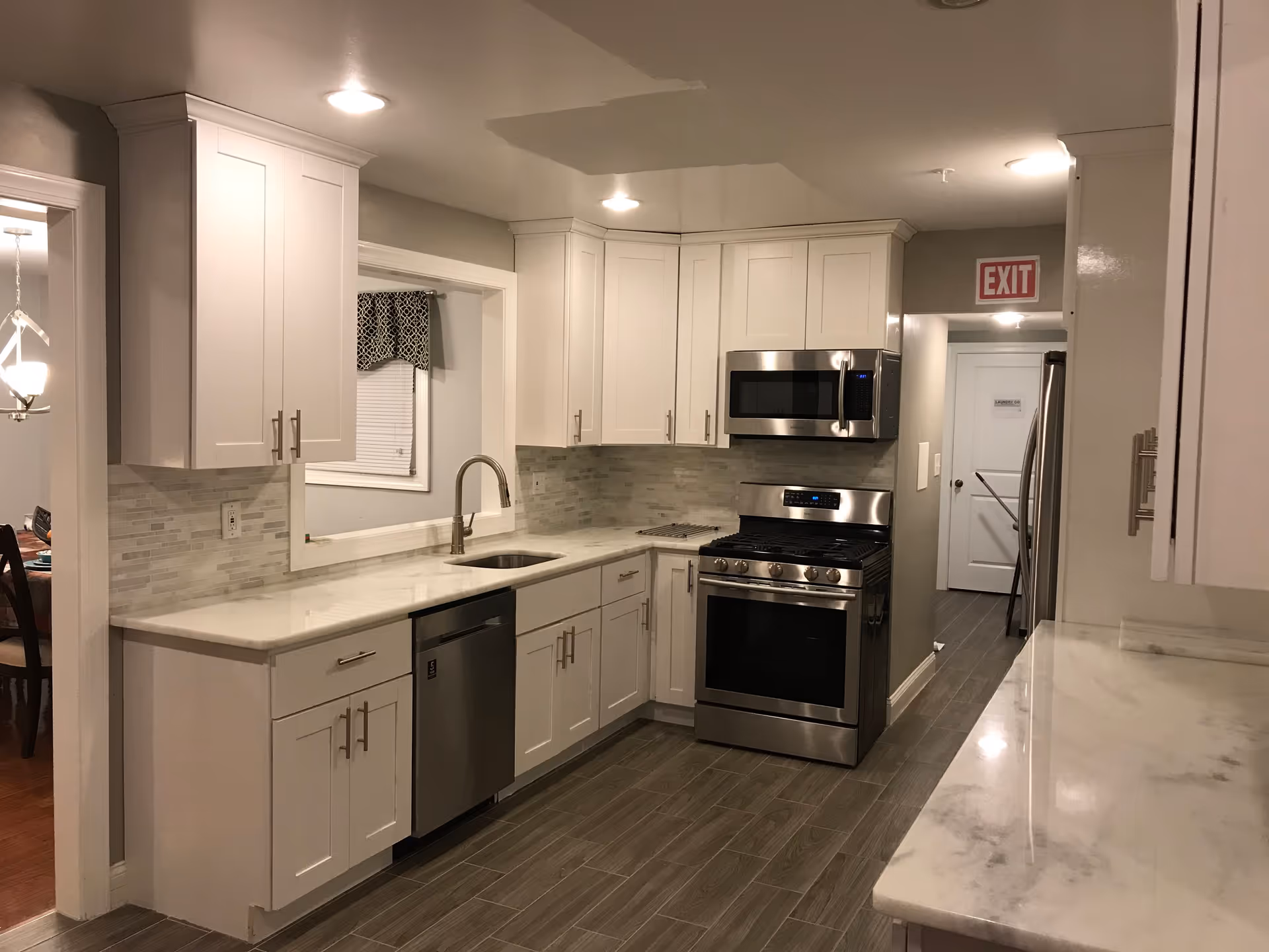 Modern kitchen with white cabinets, stainless steel appliances including a microwave, stove, and dishwasher. The kitchen has a marble countertop, a sink with a curved faucet, and a tiled backsplash. There is a window with a patterned valance above the sink and an exit sign visible in the hallway beyond the kitchen.