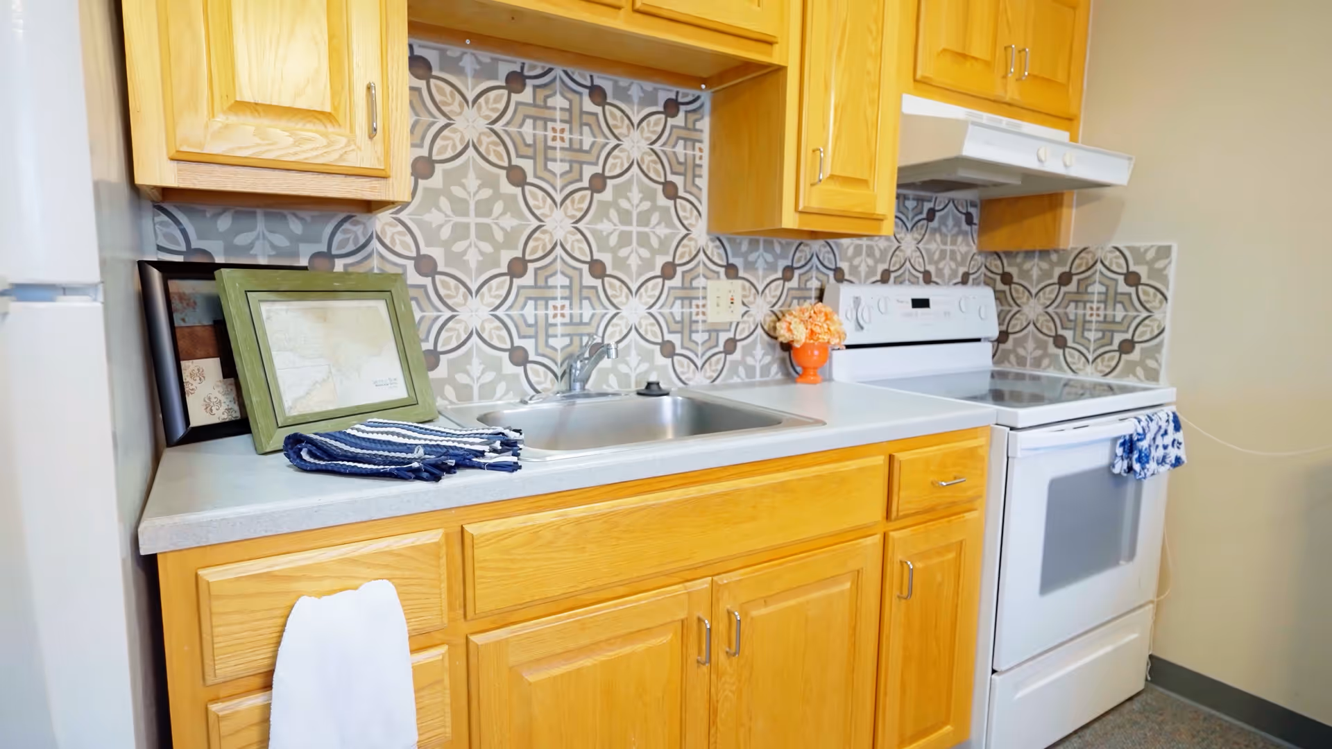 A kitchen area with light wood cabinets, a white countertop, a stainless steel sink, and a white electric stove with an oven. The backsplash features a decorative tile pattern in gray, white, and brown tones. There are framed pictures and a folded towel on the counter, and a small orange vase with flowers next to the stove.