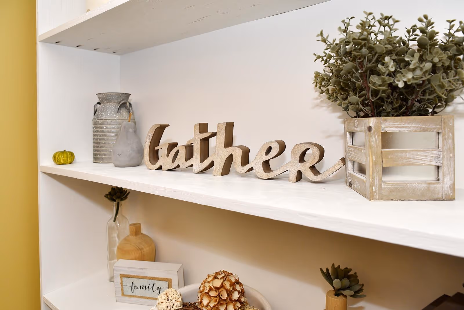 White shelving decorated with a wooden 'gather' sign, small plants, jars, and other tabletop decor.