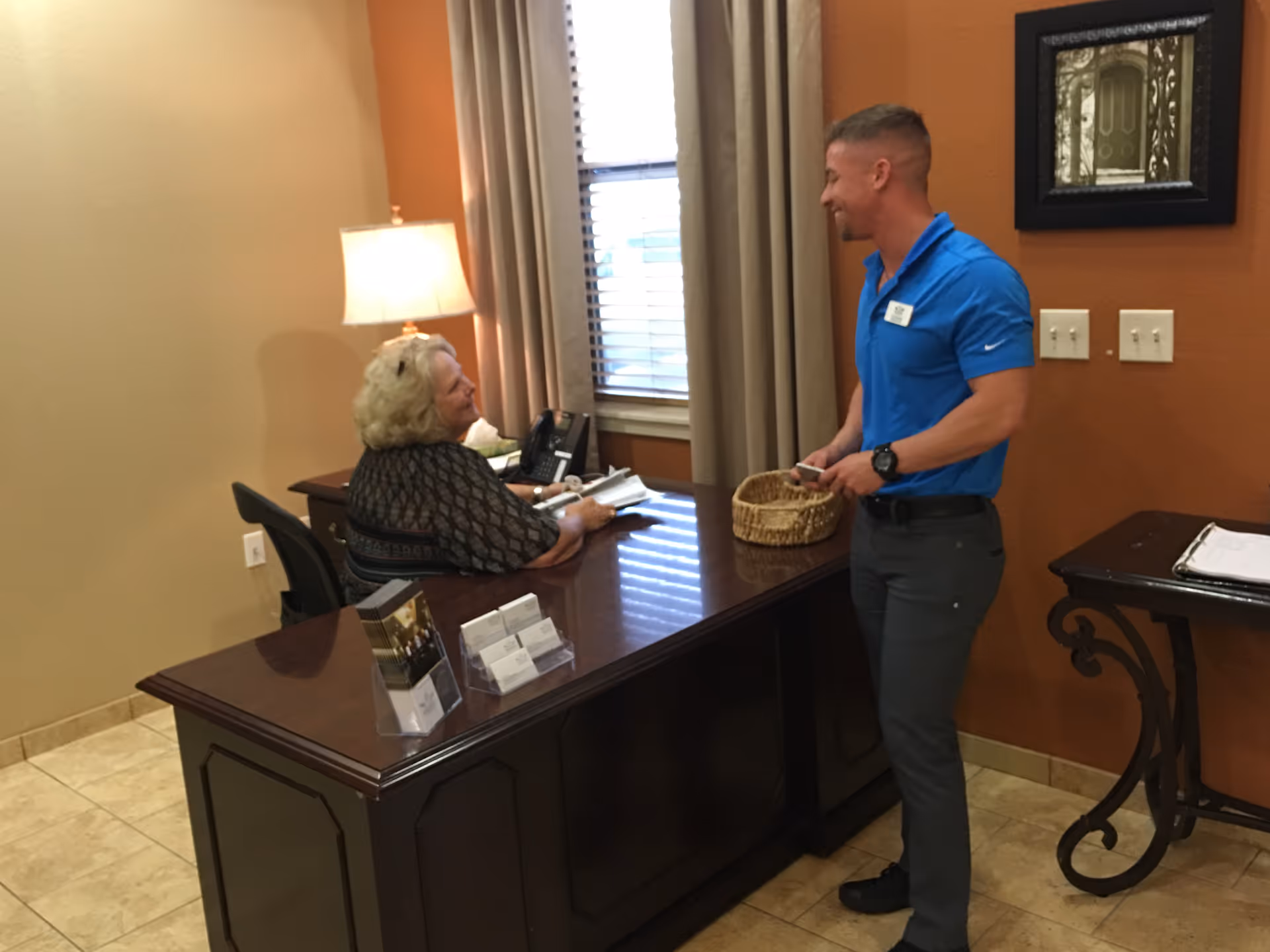 An older woman sitting behind a wooden desk in an office setting, talking to a man standing on the other side of the desk holding a basket. The room has beige walls, a window with curtains, a table lamp, and a small side table with papers on it.