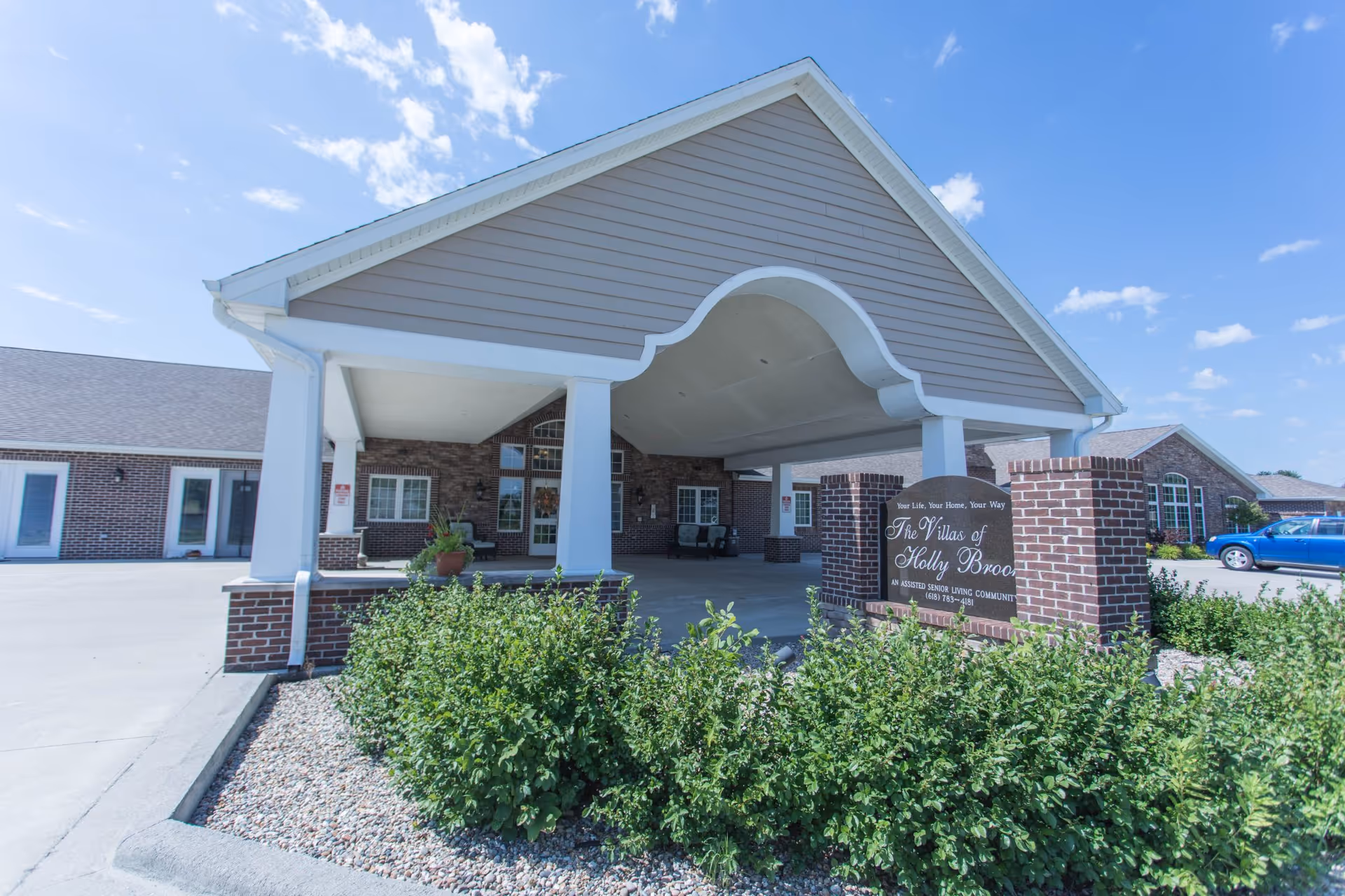 Covered drive-up entrance and canopy of The Villas of Holly Brook with brick sign and landscaping under a blue sky.