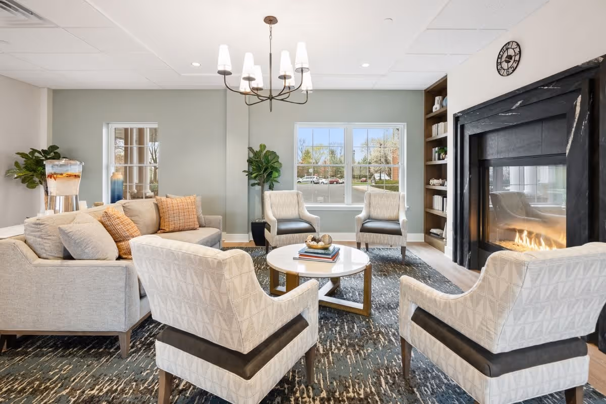 A cozy living room with a modern design featuring a beige sectional sofa with patterned cushions, four upholstered armchairs arranged around a round white coffee table with books and decorative items, a fireplace with a black mantel, built-in shelves with books and decor, a large window showing an outdoor view, a chandelier hanging from the ceiling, and a water dispenser on a side table.