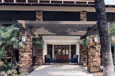 Entrance of a senior living facility with stone pillars, wooden pergola, two chairs on either side of the doorway, and palm trees surrounding the walkway.