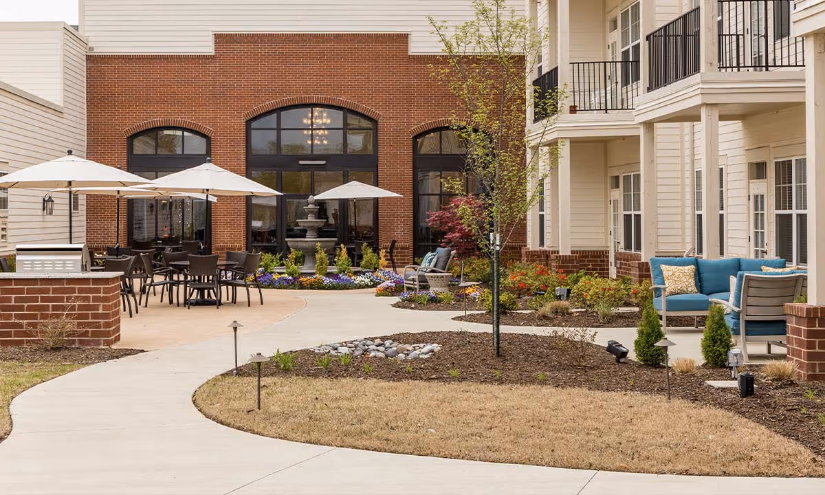 Outdoor patio area at StoryPoint Cordova featuring a curved concrete walkway, several tables with umbrellas, a water fountain, landscaped flower beds, and seating areas with blue cushioned chairs. The building exterior has brick and beige siding with balconies and large windows.
