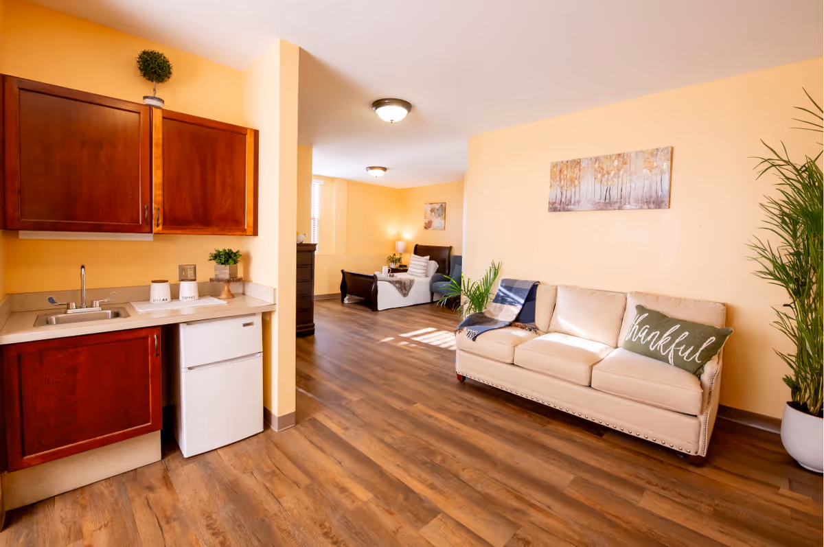 A cozy interior room with warm yellow walls and wood flooring. On the left, there is a small kitchenette with a sink, countertop, wooden cabinets, and a mini refrigerator. In the center and right, there is a beige sofa with a green pillow that says 'thankful' and a blue throw blanket. A potted plant is placed next to the sofa. In the background, there is a bed with white bedding, a nightstand with a lamp, and a dresser. The room is softly lit with ceiling lights and natural light from a window.
