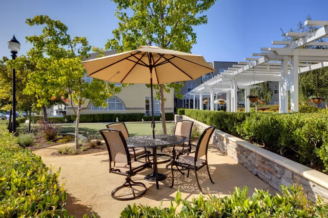 Outdoor seating area with a round metal table and four chairs under a large beige umbrella, surrounded by greenery, trees, and a stone wall with a white pergola in the background under a clear blue sky.