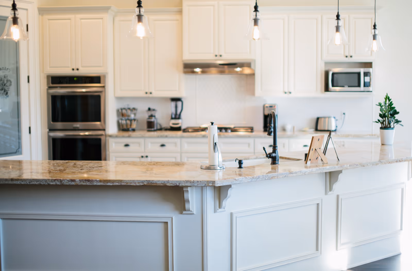 Bright and modern kitchen with white cabinetry, granite countertops, a double oven, a microwave, a toaster, a blender, a coffee maker, a paper towel holder, a black faucet, and pendant lights hanging from the ceiling.