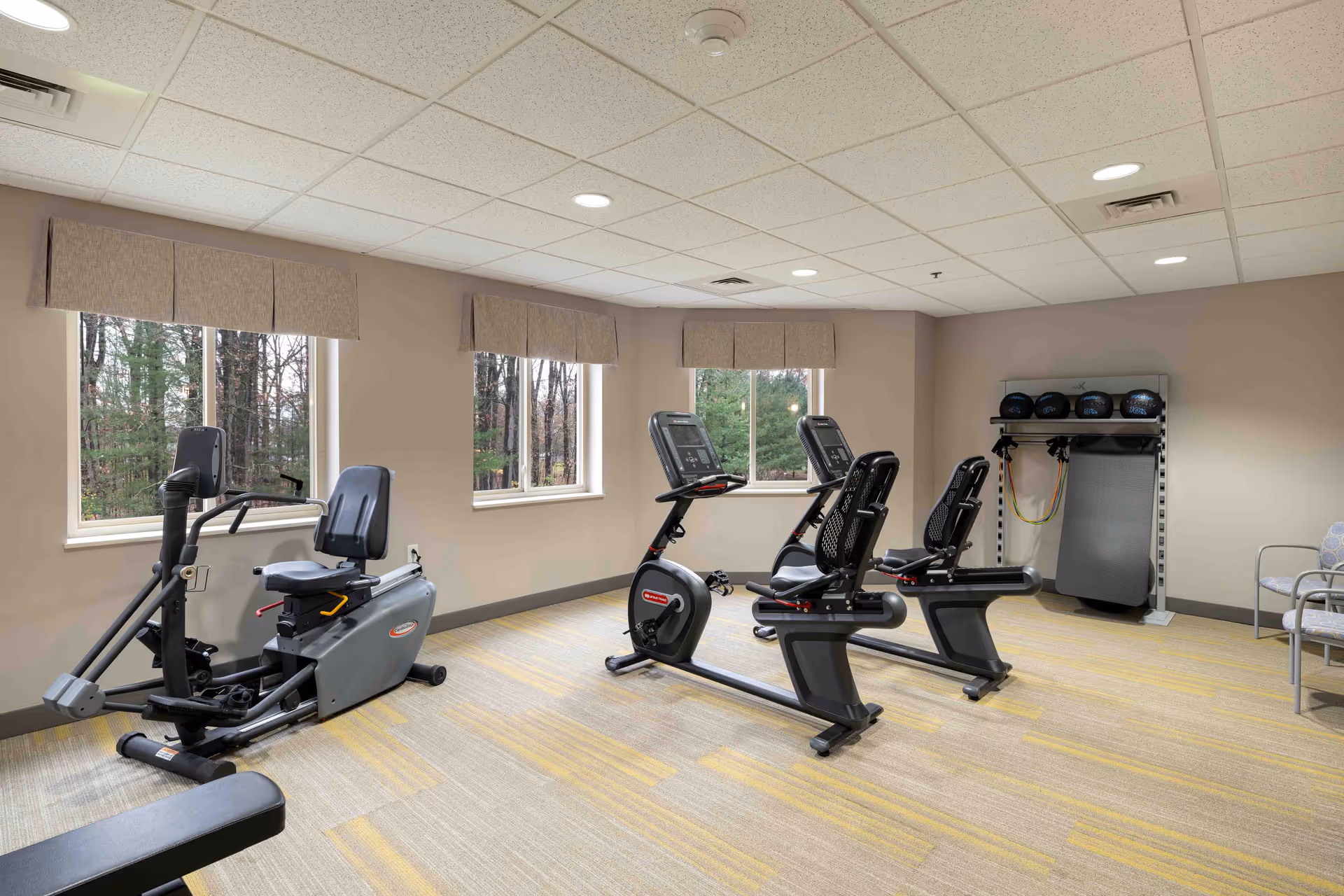 A senior living community fitness room with exercise equipment including two recumbent stationary bikes, an elliptical machine, and a rack with medicine balls and resistance bands. The room has beige walls, three windows with valances showing a wooded outdoor view, and a light-colored carpet with yellow accents.