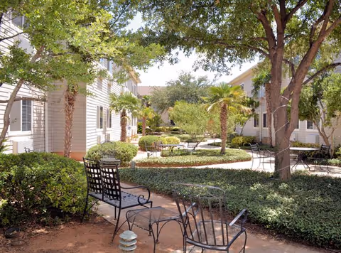 Outdoor courtyard with metal patio chairs, trees, shrubs, and winding walkways between light-colored buildings.