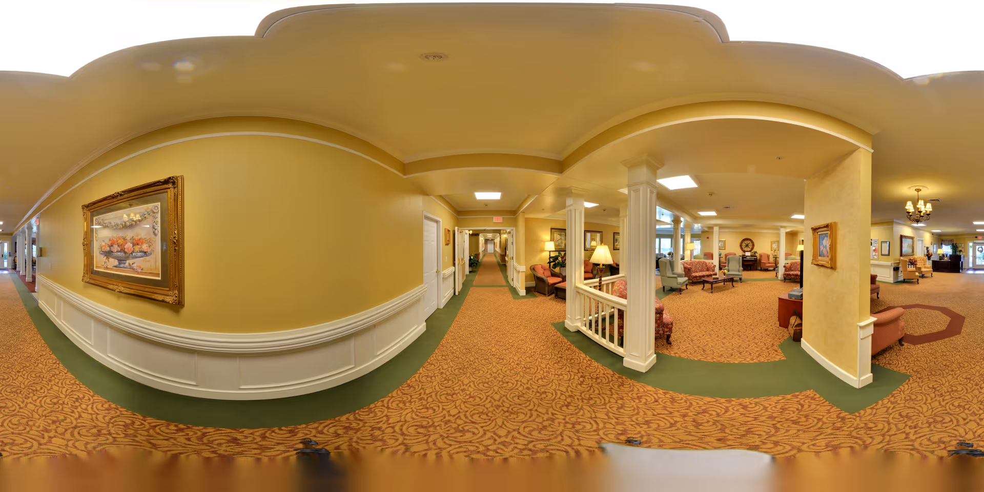 Panoramic view of a carpeted senior living hallway opening into a furnished common lounge with chairs, columns, lamps, and framed artwork.