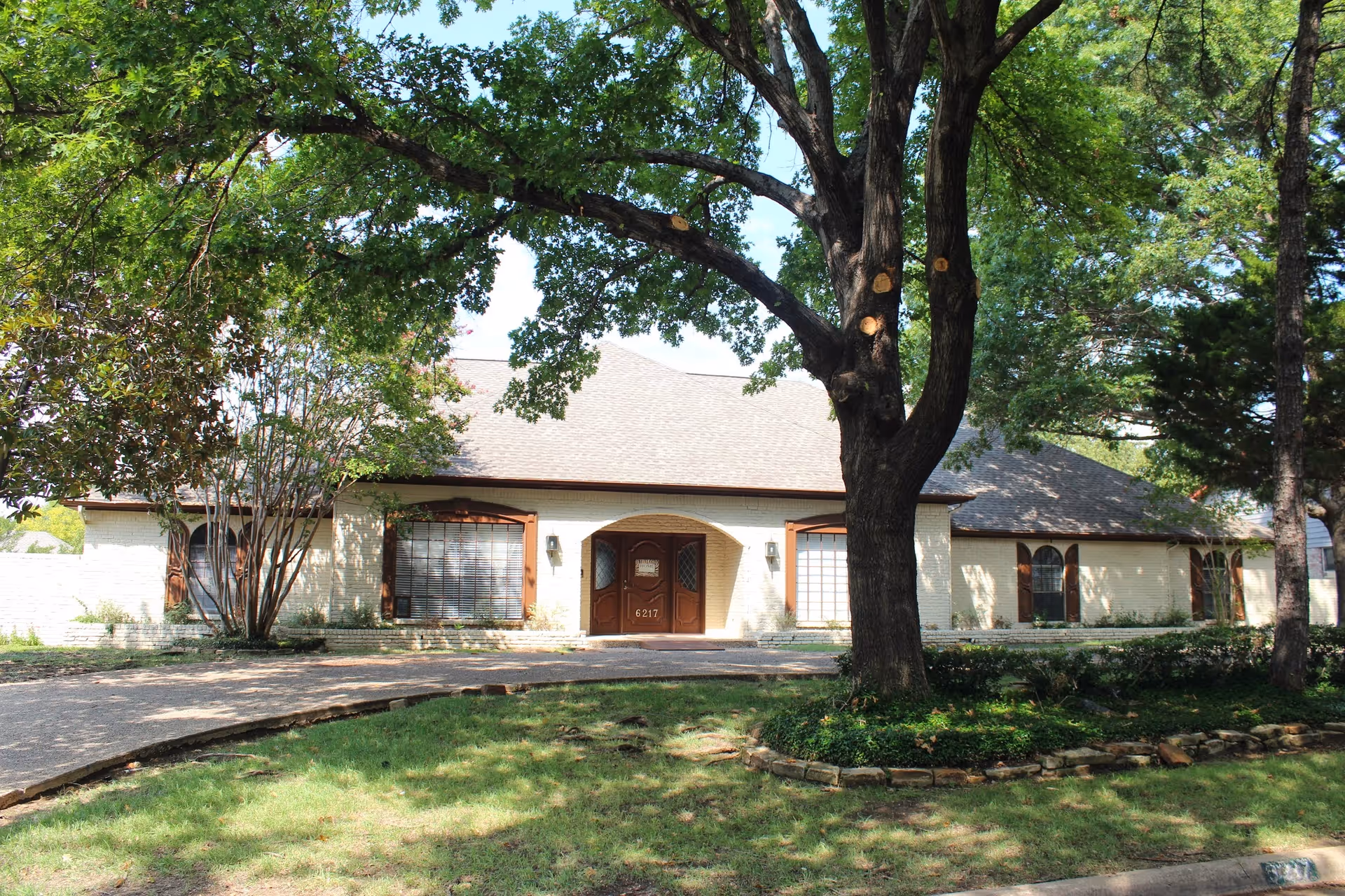 Single-story brick building with an arched central entrance, large tree in front, and a curved driveway.