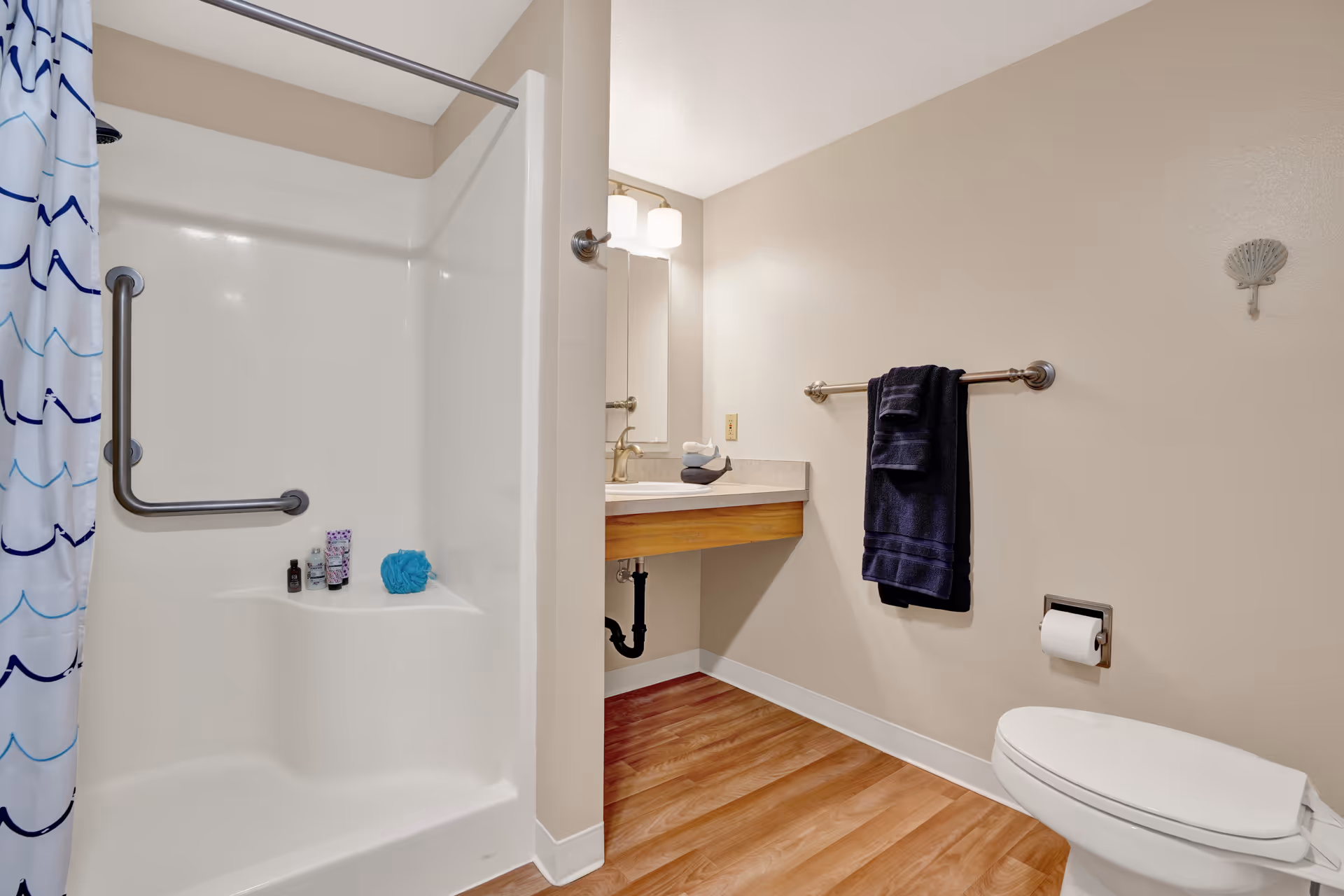 A clean bathroom with a white shower area featuring a grab bar and a shower curtain with blue wave patterns. The bathroom has a wooden floor, a wall-mounted sink with a mirror and light above it, a towel rack with a dark blue towel, a toilet with a toilet paper holder, and a small decorative shell on the wall.
