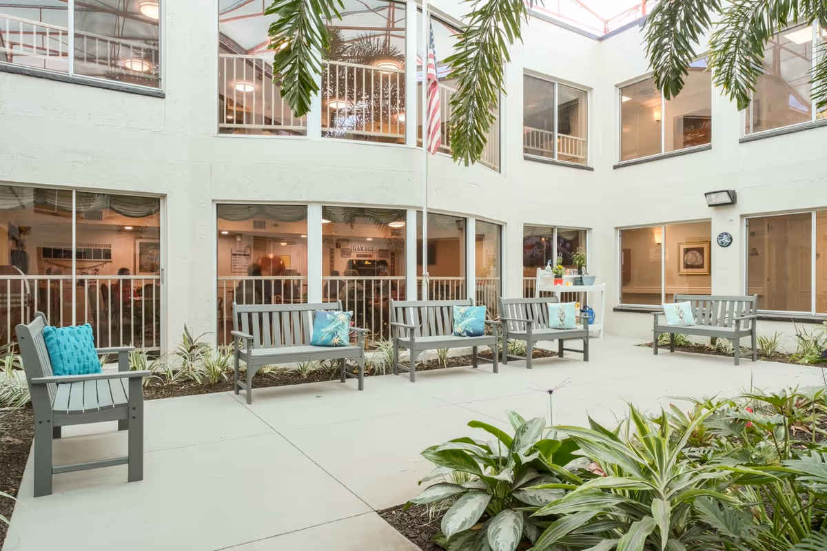 A bright indoor courtyard with several gray wooden benches adorned with colorful cushions. The courtyard is surrounded by large windows showing interior rooms with people and decor. There are plants and greenery along the edges and a flagpole with an American flag in the center.