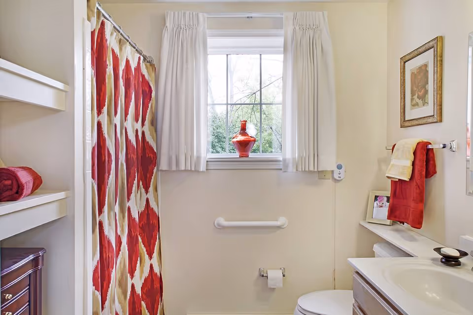 Bright bathroom with a red-and-white patterned shower curtain, a window with a red vase, sink, toilet, grab bar, and towel rack.