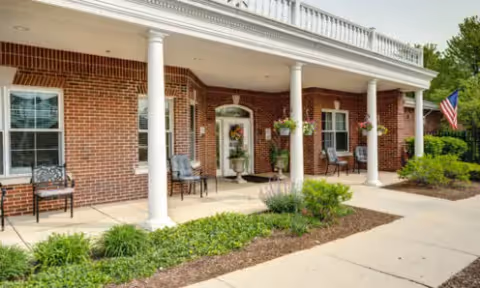 Front exterior view of a brick building with white columns supporting a porch roof. There are chairs and hanging flower baskets on the porch, an American flag on the right side, and landscaping with bushes and plants along the sidewalk.