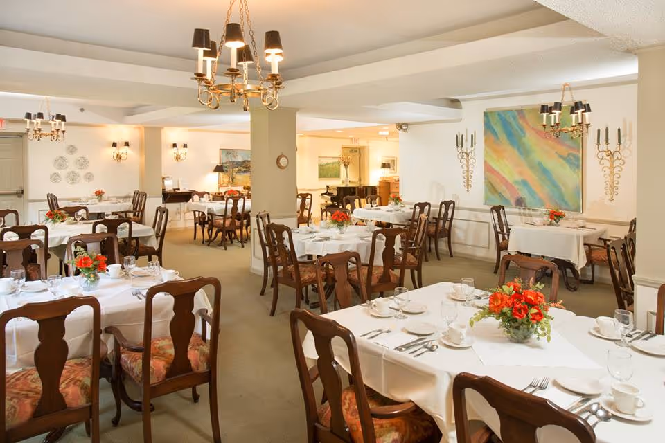 A well-lit dining room with multiple tables covered in white tablecloths, each set with cups, glasses, plates, and silverware. Wooden chairs with patterned cushions surround the tables. The room features chandeliers, wall sconces, colorful artwork, and floral centerpieces on each table.