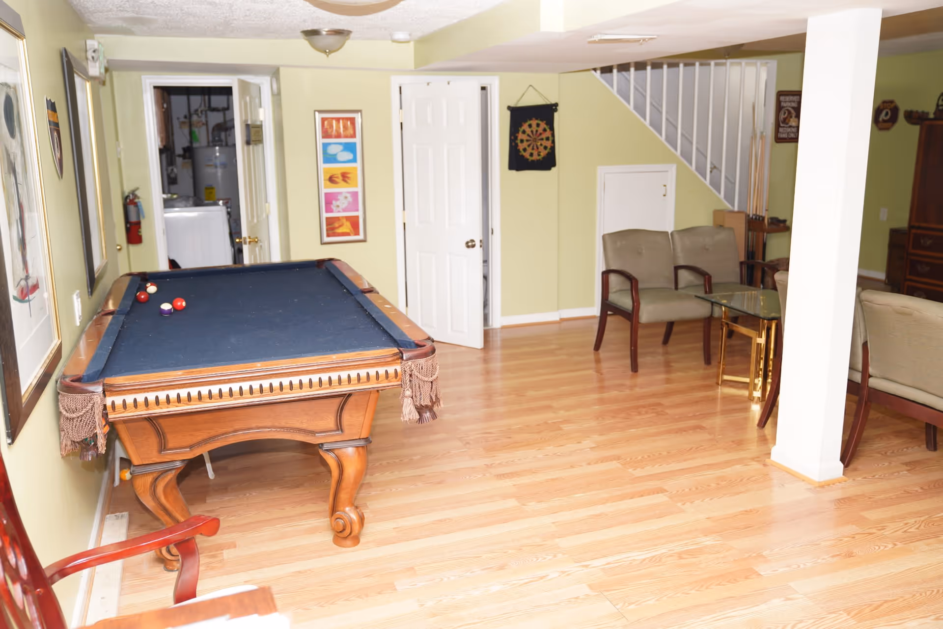 A recreational room with a pool table featuring a dark blue felt surface and a few billiard balls on it. The room has light wooden flooring and light green walls. There are two beige armchairs with wooden armrests and a glass-top side table near a white column. A dartboard hangs on the wall near a staircase with white railings. The room also has framed artwork on the walls and an open door leading to another room with a washing machine and water heater visible.