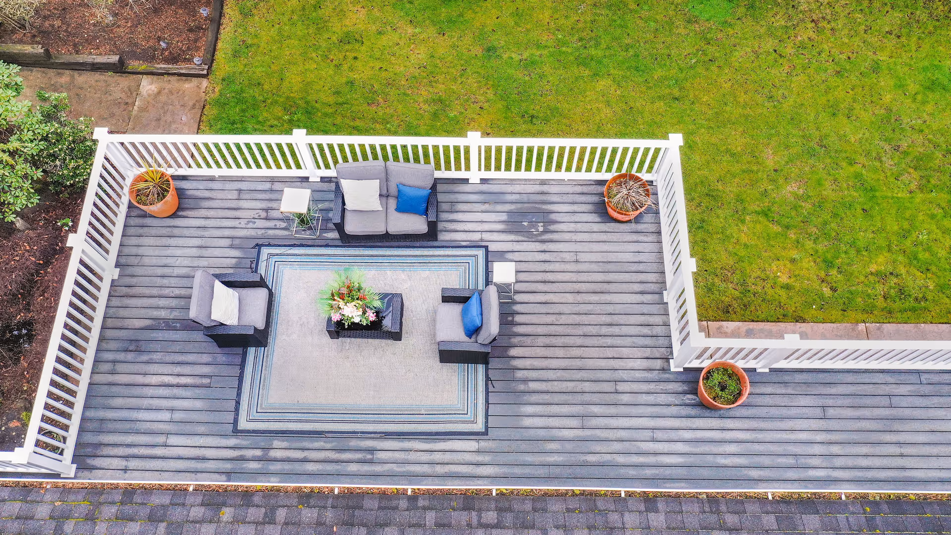 Aerial view of a wooden deck with outdoor seating including a loveseat and two armchairs with cushions, a small table, a rectangular rug, and three potted plants. The deck is surrounded by a white railing and adjacent to a grassy lawn.