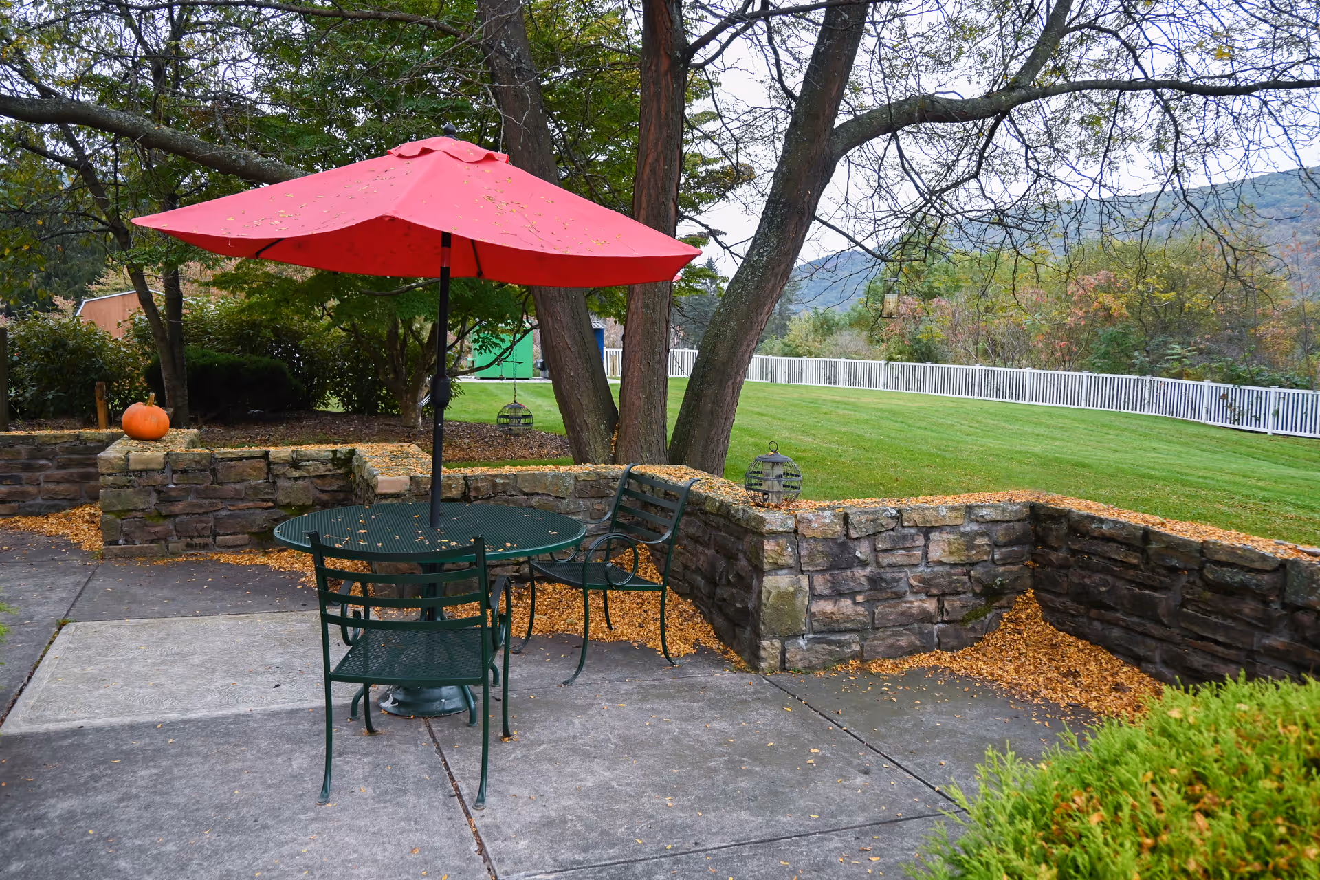 Outdoor patio area with a round metal table and four chairs under a red umbrella. The patio is surrounded by a low stone wall with some fallen leaves scattered on the ground. In the background, there is a grassy lawn, trees, and a white fence with hills visible in the distance.