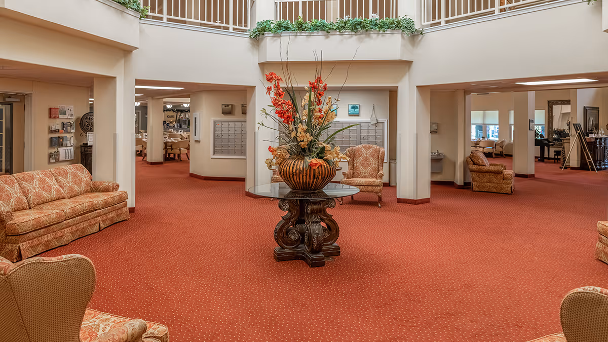 Spacious senior living facility lobby with red carpet, upholstered seating, and a central table topped with a large floral arrangement.