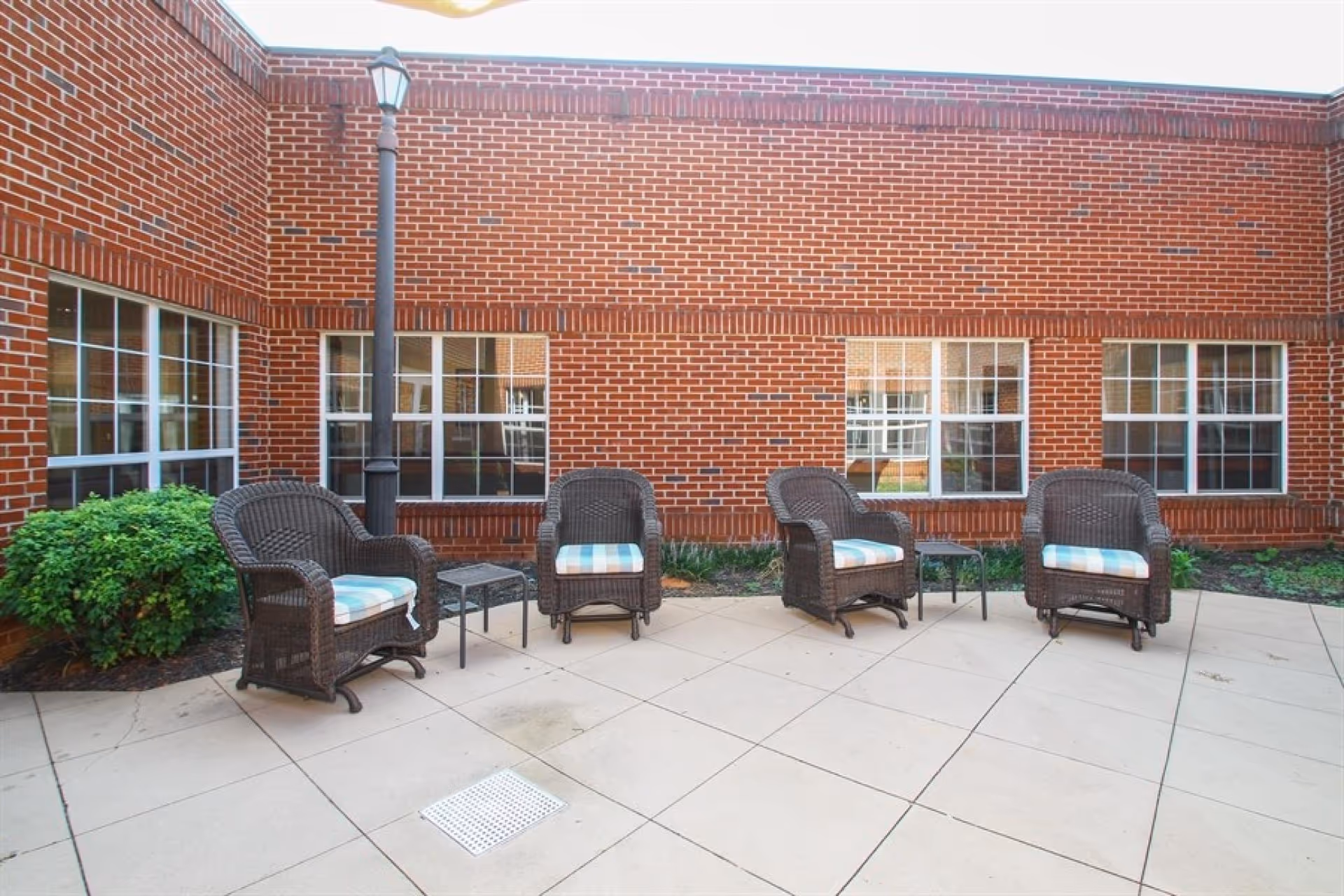 Outdoor patio area with four wicker chairs with blue and white cushions arranged around two small tables, set against a red brick building with multiple windows and a black lamppost.