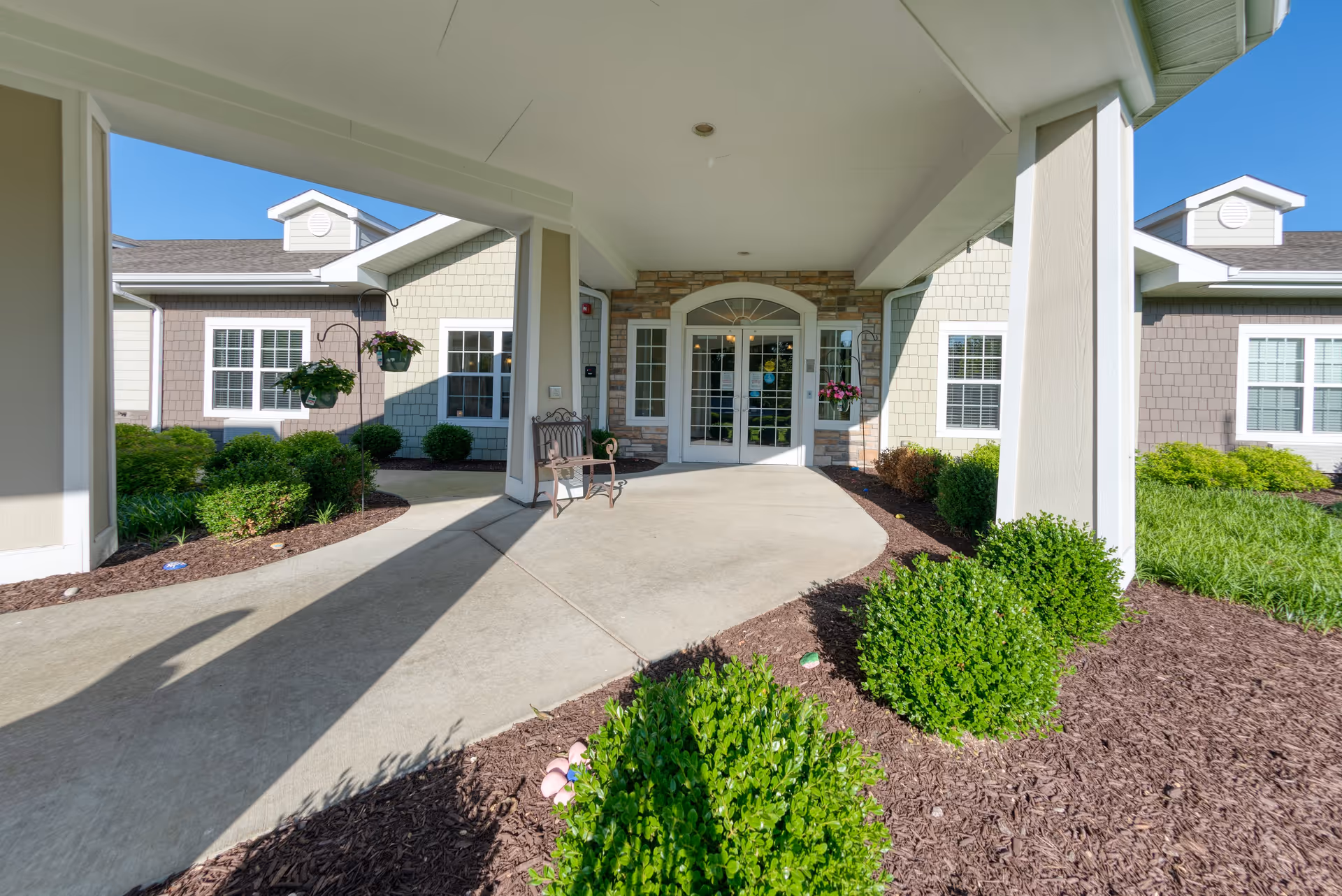 Covered entrance area of a senior living facility with a concrete walkway, green shrubs, hanging flower pots, and a bench near the double glass doors leading inside. The building exterior features light-colored siding and stone accents under a clear blue sky.
