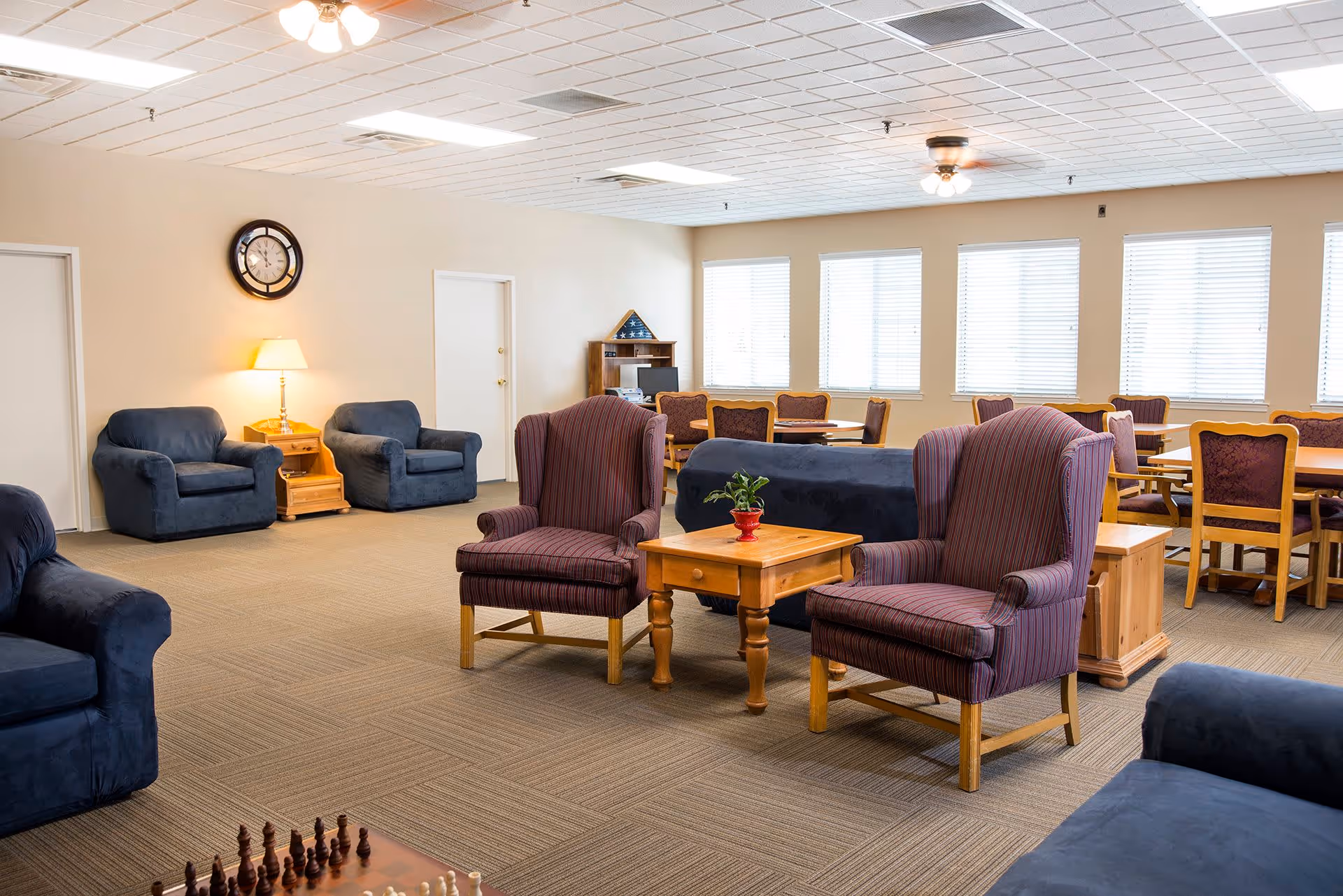 A spacious living room area in a senior living facility with multiple upholstered chairs and sofas arranged around wooden tables. There is a chessboard in the foreground, a clock and a lamp on a side table against the wall, and several windows with blinds letting in natural light. In the background, there are dining tables and chairs near the windows.