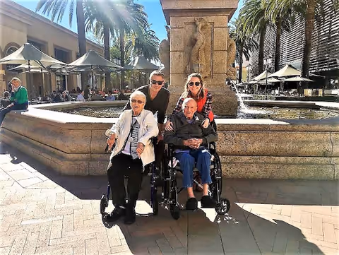 Two elderly individuals in wheelchairs are positioned in front of a large stone fountain with palm trees and outdoor seating areas in the background. Two younger adults stand behind them, smiling. The setting appears to be a sunny outdoor plaza or courtyard.