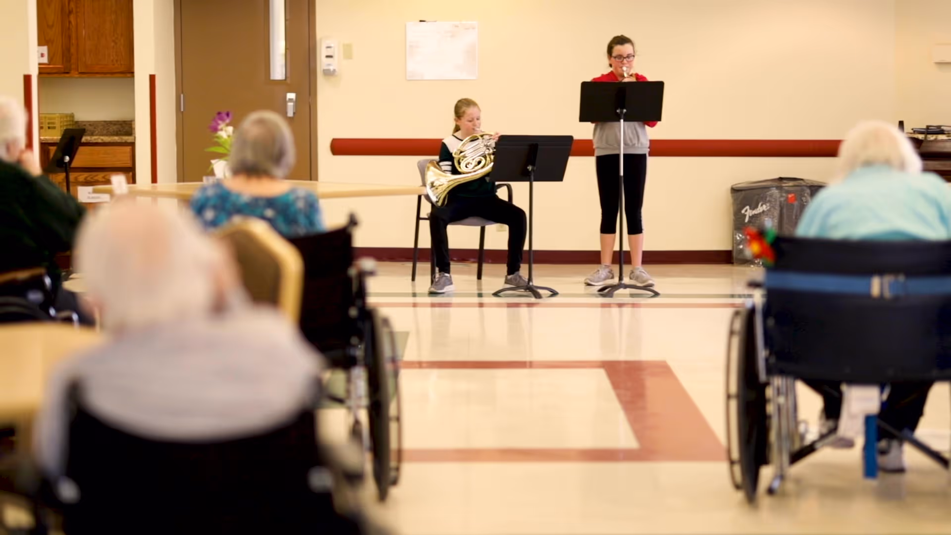 Two young musicians perform at the front of a senior facility activity room while elderly residents in wheelchairs watch.