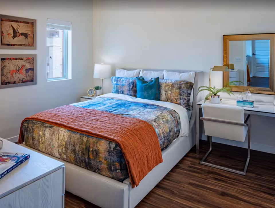 A neatly made bed with a colorful patterned bedspread and an orange throw blanket in a bright bedroom. There are three decorative pillows on the bed. To the right of the bed is a white desk with a gold-framed mirror, a white chair, a small plant, and a lamp. On the left side of the bed is a nightstand with a lamp, a small clock, and a plant. Two framed artworks hang on the wall next to a window with a white blind. The floor is wooden.
