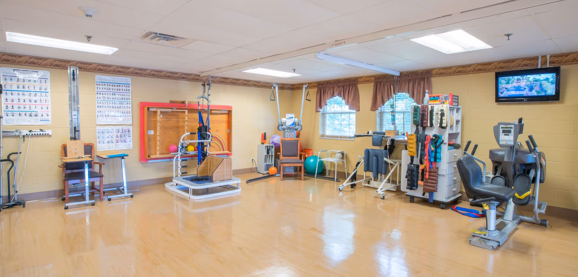 A bright rehabilitation/exercise room with various therapy equipment, chairs, and exercise machines.